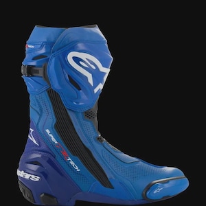 Könnte beinhalten: Ein blauer Alpinestars Supertech Motorrad-Rennstiefel mit weißem Logo auf dem Schienbeinschutz, schwarzen Seitenteilen, perforierten Lederbereichen, einer verstärkten Fersenkappe mit dem Wort stars und einem Zehenschleifer an der Vorderseite.
