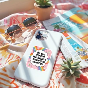 May include: White smartphone with a sticker that reads "Be the person Mr. Rogers knew you could be" in a cloud-shaped design with a rainbow and flowers. Gold-rimmed sunglasses and a magazine are in the background.