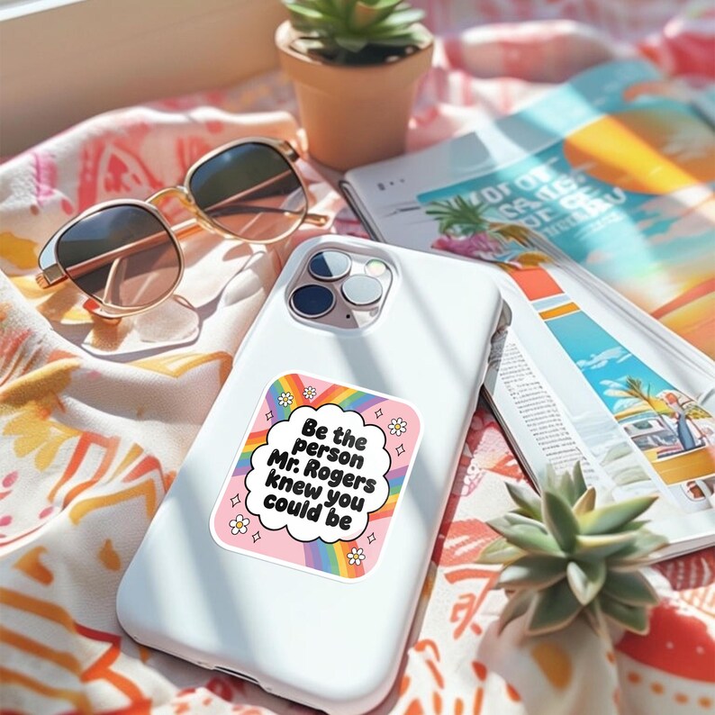May include: White smartphone with a sticker that reads "Be the person Mr. Rogers knew you could be" in a cloud-shaped design with a rainbow and flowers. Gold-rimmed sunglasses and a magazine are in the background.