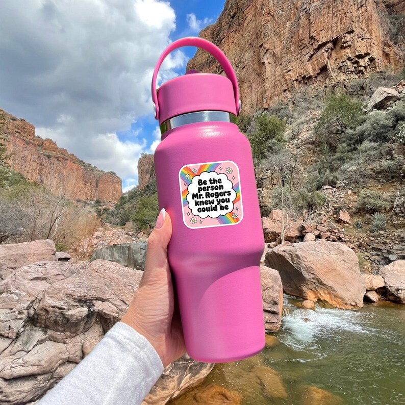 May include: A pink insulated water bottle with a matching handle and lid. A sticker on the bottle reads "Be the person Mr. Rogers knew you could be" in a rainbow design. The bottle is held in front of a scenic outdoor background.