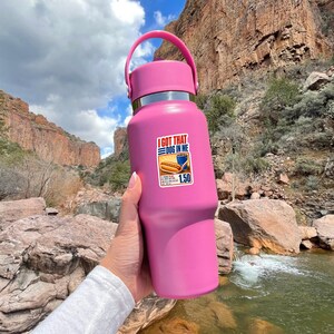 Puede incluir: Una botella de agua aislada de color rosa mate con un asa en la tapa. Una pegatina rectangular en la parte frontal muestra un perrito caliente y una bebida azul con el texto I GOT THAT DOG IN ME y 1.50, referente a un men&uacute; de comida.