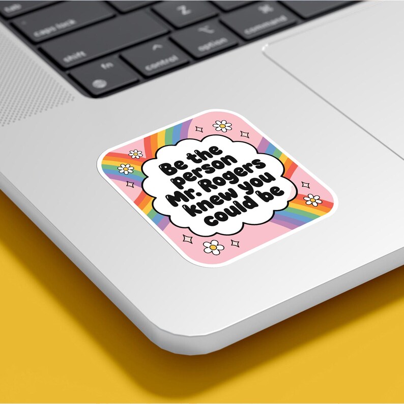 May include: A square sticker with a pink background and a rainbow border. The sticker has the text "Be the person Mr. Rogers knew you could be" in a white cloud-shaped text box. Small daisy and diamond shapes surround the text.