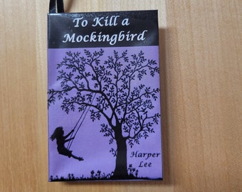 May include: A purple book with a black ribbon bookmark. The book cover features a silhouette of a girl on a swing in a tree. The title "To Kill a Mockingbird" and the author "Harper Lee" are printed in black on the cover.