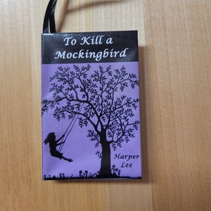 May include: A purple book with a black ribbon bookmark. The book cover features a silhouette of a girl on a swing in a tree. The title "To Kill a Mockingbird" and the author "Harper Lee" are printed in black on the cover.