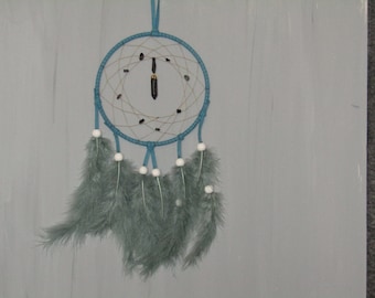 May include: A blue dream catcher with white beads and teal feathers hanging from it. The dream catcher has a black crystal hanging in the center.