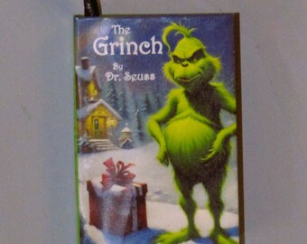 May include: A rectangular ornament featuring the Grinch from Dr. Seuss's "The Grinch." The Grinch is depicted in his signature green color, standing in a snowy scene with a gift box. The ornament has the title "The Grinch" and the author's name.