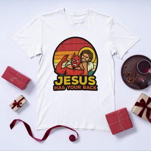 Könnte beinhalten: Weißes T-Shirt mit einer Grafik, die Jesus beim Ringen mit einem roten Teufel vor einem Hintergrund im Sonnenuntergangsstil zeigt. Der Text "JESUS HAS YOUR BACK" ist unter dem Bild aufgedruckt. Das T-Shirt ist von verpackten Geschenken und einem dekorativen Teller umgeben.