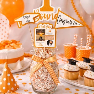 Puede incluir: Una escena de fiesta de graduación con decoraciones naranjas y blancas. Un tarro lleno de papel triturado y una cinta es la pieza central, rodeado de cupcakes, globos y letreros que dicen "Congrats Lils" y "Class of 2023".