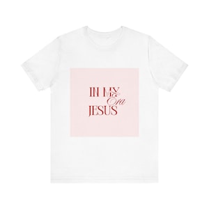 May include: White t-shirt featuring the burgundy text "IN MY Ora JESUS" on a light pink square. The text is in a script font. The shirt is a short-sleeved, casual style.