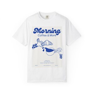 Op de afbeelding: Wit t-shirt met een blauwe afbeelding met de tekst "Morning Coffee & More". De afbeelding toont een croissant, koffie die wordt geschonken en de tekst "Coffee first. Chill vibes only. Golden mornings, cozy moments."