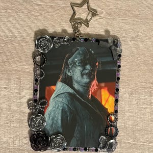 May include: A rectangular photo frame with a picture of a person wearing a mask. The frame is decorated with black, silver, and purple embellishments, including roses, beads, and bows. A silver star-shaped keychain is attached.