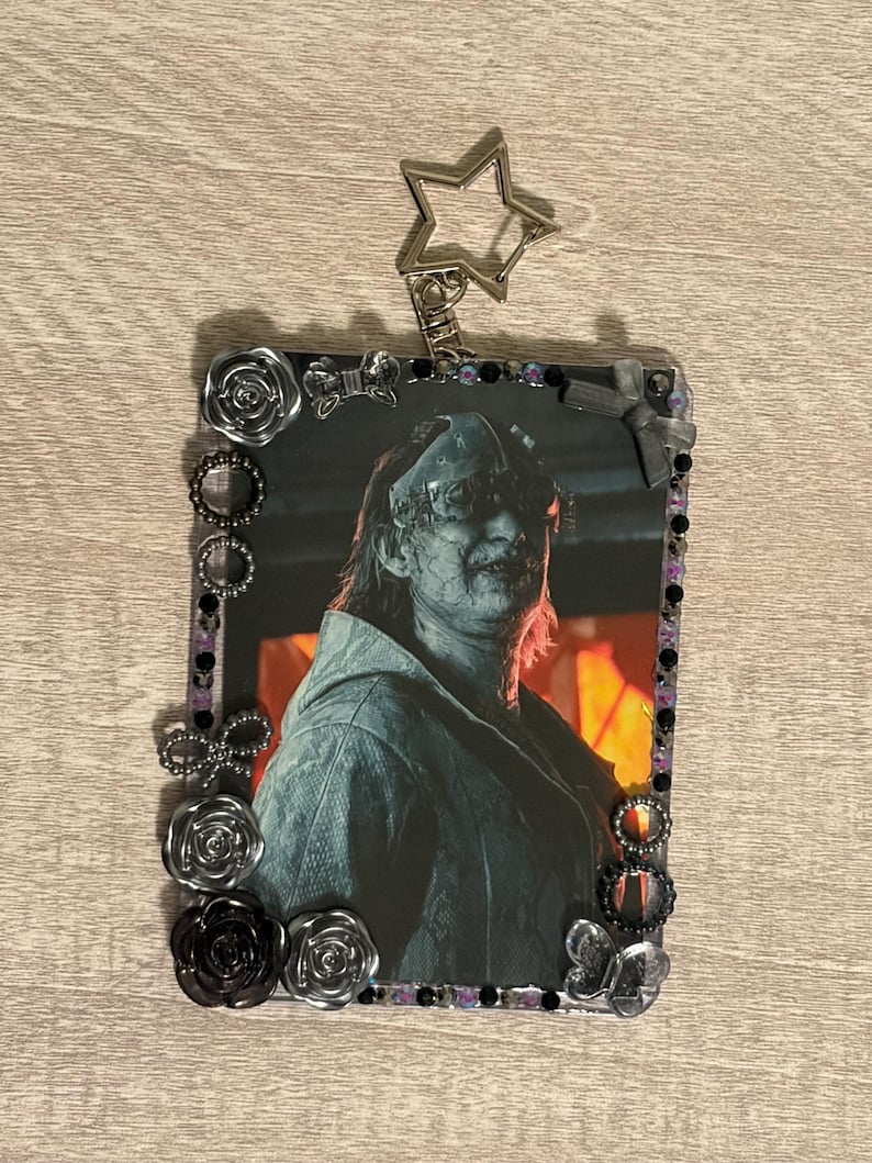 May include: A rectangular photo frame with a picture of a person wearing a mask. The frame is decorated with black, silver, and purple embellishments, including roses, beads, and bows. A silver star-shaped keychain is attached.
