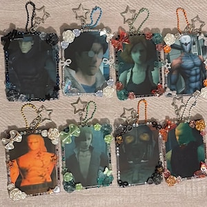 May include: Eight rectangular keychains featuring character portraits from the Metal Gear Solid video game series. Each frame is decorated with small plastic flowers, bows, butterflies, and beads in various colours, attached to a ball chain with a star charm.