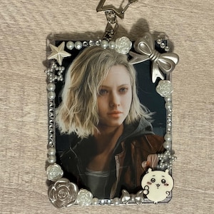 May include: A rectangular keychain featuring a portrait of a person with blonde hair. The frame is decorated with pearls, silver bows, and star-shaped charms. A small, white cartoon character is attached to the bottom right corner, and a star-shaped clasp is at the top.