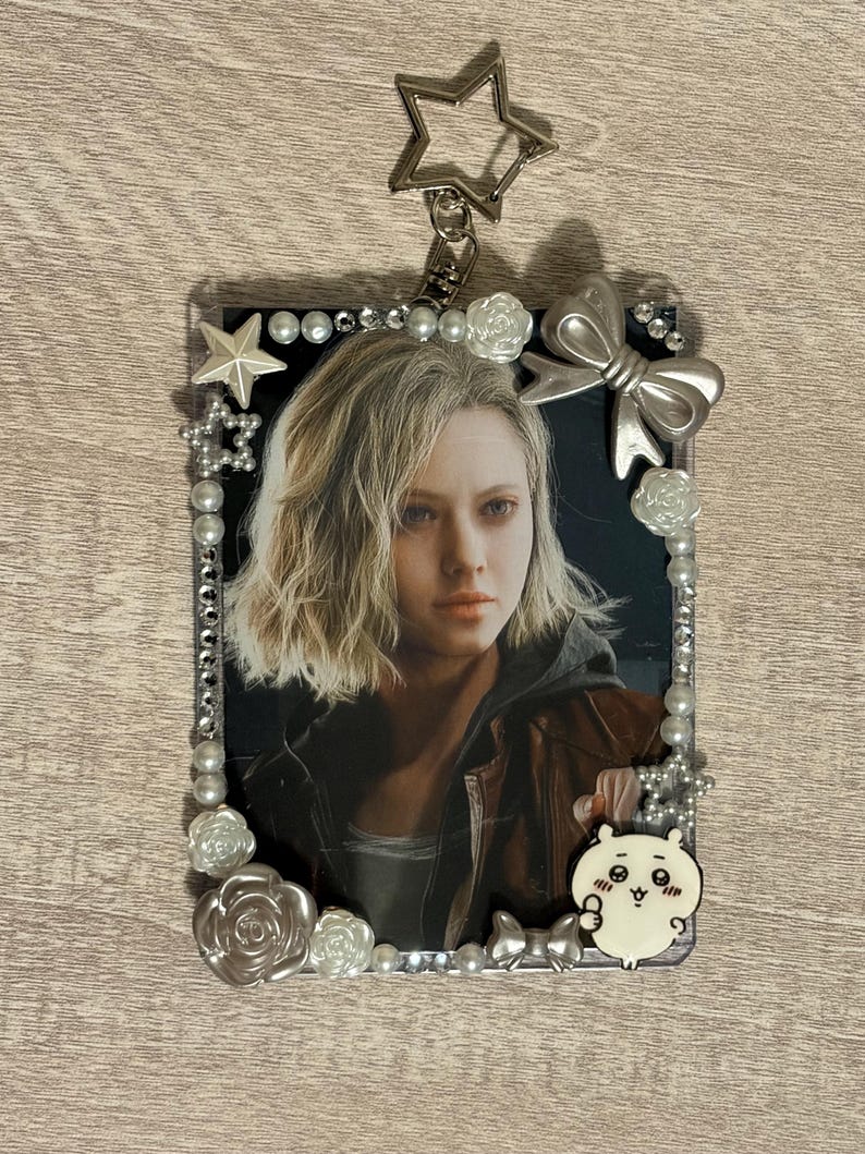 May include: A rectangular keychain featuring a portrait of a person with blonde hair. The frame is decorated with pearls, silver bows, and star-shaped charms. A small, white cartoon character is attached to the bottom right corner, and a star-shaped clasp is at the top.