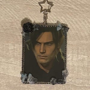 May include: A rectangular keychain featuring a portrait of a person with dark hair and a serious expression. The image is framed with a silver border and adorned with small black roses and blue butterfly accents. A silver star-shaped charm hangs from the top.