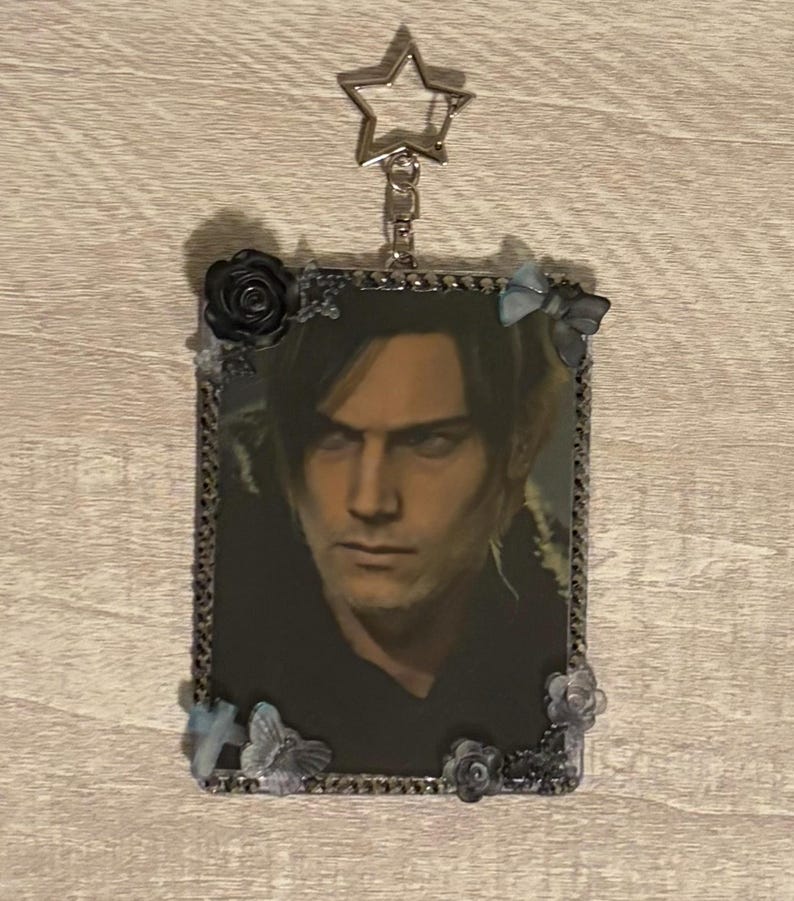 May include: A rectangular keychain featuring a portrait of a person with dark hair and a serious expression. The image is framed with a silver border and adorned with small black roses and blue butterfly accents. A silver star-shaped charm hangs from the top.