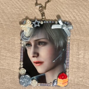 May include: A rectangular keychain featuring a portrait of a person with short, light-colored hair. The image is framed with pearls, bows, and floral accents. A star-shaped charm and a lobster clasp are attached at the top. A small cake-shaped charm is at the bottom.