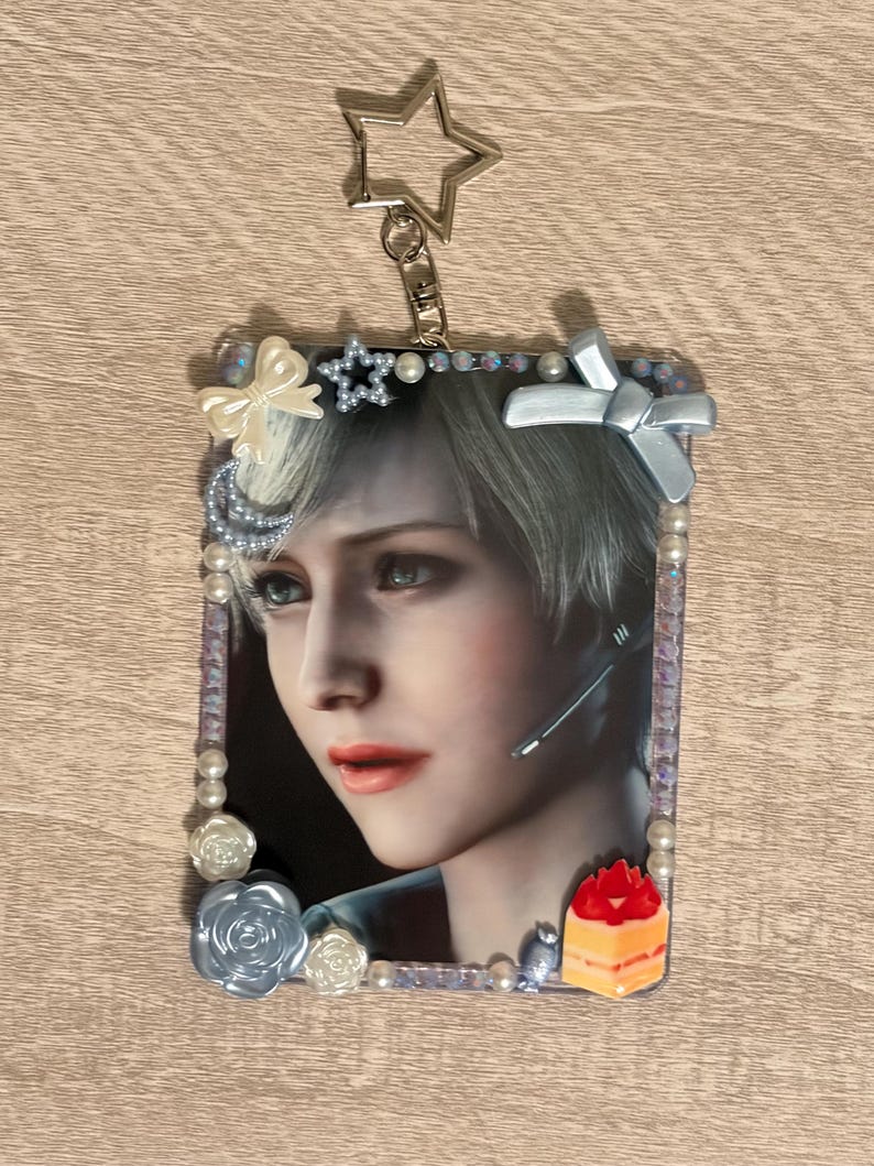 May include: A rectangular keychain featuring a portrait of a person with short, light-colored hair. The image is framed with pearls, bows, and floral accents. A star-shaped charm and a lobster clasp are attached at the top. A small cake-shaped charm is at the bottom.