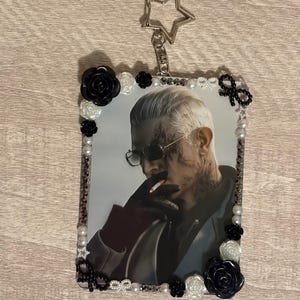 May include: A rectangular keychain featuring a portrait of a person with sunglasses and tattoos, surrounded by a decorative border of pearls, black and white roses, and bows. A silver star-shaped charm is attached to the top.