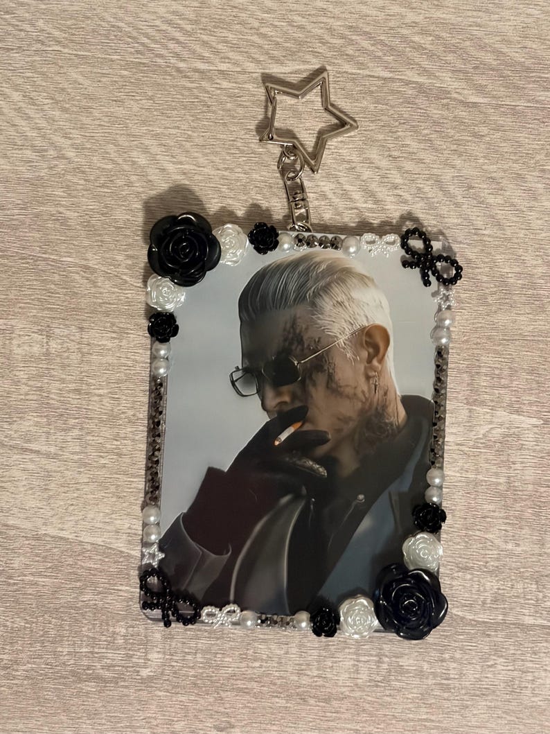 May include: A rectangular keychain featuring a portrait of a person with sunglasses and tattoos, surrounded by a decorative border of pearls, black and white roses, and bows. A silver star-shaped charm is attached to the top.