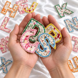 May include: A collection of fabric letter patches featuring various floral patterns on a cream background. Each letter is outlined with a thick, vibrant thread border in colors including pink, blue, green, yellow, and orange.