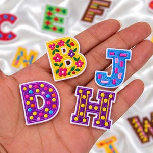 May include: Four embroidered alphabet letter patches in the letters B, J, D, and H. The B is yellow with pink and purple flowers. The J is blue with pink rectangles. The D is purple with colorful dots. The H is purple with yellow dots.