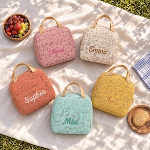 May include: Five square lunch bags with top handles and a leopard print pattern in pink, beige, orange, mint green, and yellow. Each bag features a front pocket and is embroidered with a name: Olivia, Emma, Sophia, Mia, or Ava in matching thread.