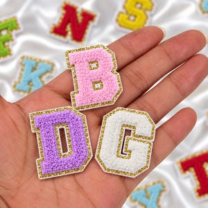 May include: Three varsity-style letter patches featuring a textured pink B, a purple D, and a white G. Each letter has a thick, glittery gold border. These embroidered patches are designed for fabric application and display a raised, fuzzy surface.