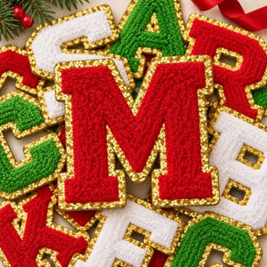 May include: A collection of plush, textured letter patches in red, green, and white, each outlined with a shimmering gold sequin border. The letters are arranged in a layered pile, featuring a prominent red letter M in the center.
