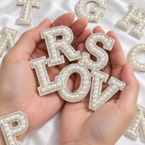 May include: A collection of alphabet letter patches featuring white faux pearls and clear rhinestones set in a silver-toned border. The letters R, L, O, V, and S are prominently displayed, surrounded by other scattered letter patches.