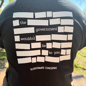 Peut inclure: T-shirt noir avec texte blanc disposé en forme rectangulaire. Le texte dit "the government wouldn't lie to you" et "ILLEGITIMATE CONCERNS" en dessous. Le t-shirt est fait d'une matière douce.