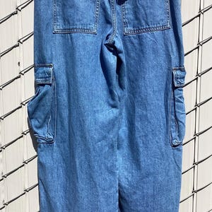 May include: Light blue denim cargo pants with multiple pockets. The pants have two large pockets on the back and two cargo pockets on the sides. The denim is a medium wash, and the pants appear to be new.