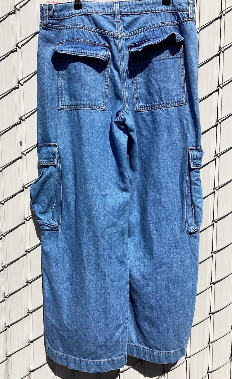 May include: Light blue denim cargo pants with multiple pockets. The pants have two large pockets on the back and two cargo pockets on the sides. The denim is a medium wash, and the pants appear to be new.