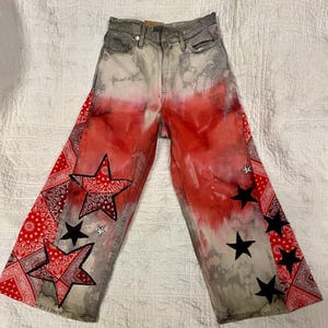 Op de afbeelding: Verontreinigde denim jeans met een rood-wit tie-dye effect. De jeans hebben rode bandana stoffen panelen met ster applicaties in rood en zwart. De jeans hebben een vervaagde grijze wassing en een klassiek vijf-zakken ontwerp.