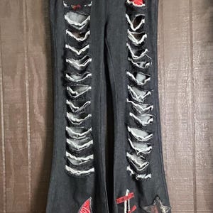 May include: Black denim jeans featuring extensive vertical distressing along the front legs with frayed edges. The pants include a red bandana-print star patch, a floral rose appliqué, a small star patch, and red fabric accents on the thighs.