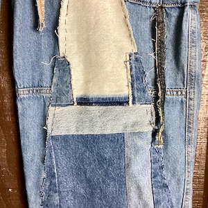 May include: Close-up of a section of denim fabric with patchwork details. The design features various shades of blue denim, with visible stitching and a contrasting light-colored fabric insert. The overall aesthetic is a distressed, handcrafted style.