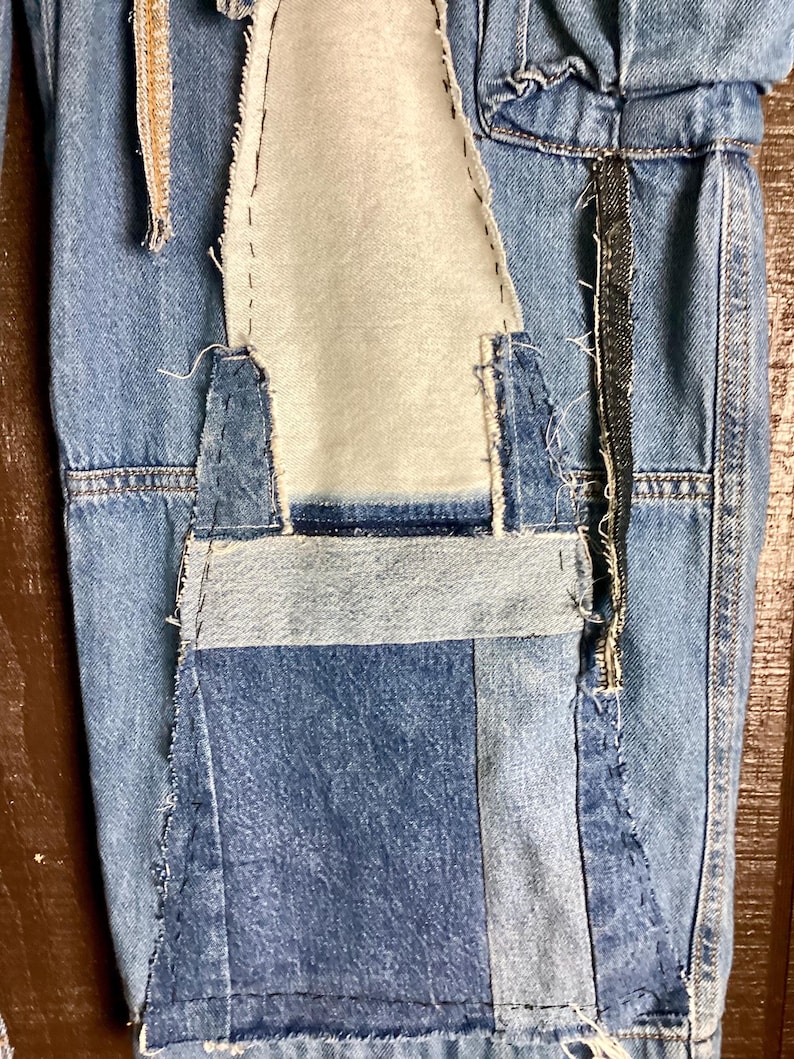 May include: Close-up of a section of denim fabric with patchwork details. The design features various shades of blue denim, with visible stitching and a contrasting light-colored fabric insert. The overall aesthetic is a distressed, handcrafted style.