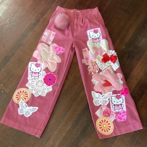 May include: Pink wide-leg pants featuring various appliqués including Hello Kitty characters, white lace doilies, fabric flowers, butterflies, a red bow, and a fuzzy pink pom-pom attached near the waistband. The pants have a button closure.