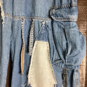 May include: Close-up of a pair of light blue denim pants with patchwork and distressed details. The pants feature a unique design with various denim patches and visible stitching, creating a textured and artistic look. The design includes a white fabric patch.