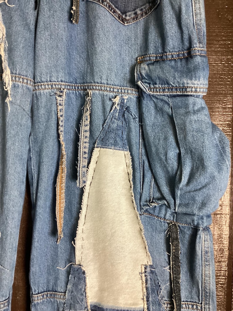 May include: Close-up of a pair of light blue denim pants with patchwork and distressed details. The pants feature a unique design with various denim patches and visible stitching, creating a textured and artistic look. The design includes a white fabric patch.