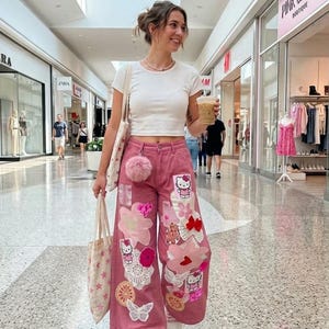 May include: A white short-sleeved crew neck t-shirt paired with wide-leg pink denim pants featuring various patches including Hello Kitty characters, flowers, butterflies, and lace accents. A fluffy pink pom-pom hangs from the waistband.