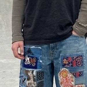 May include: A black short-sleeved t-shirt layered over a brown and black striped long-sleeved shirt, paired with blue denim jeans featuring various colorful patches, including a large skull design, floral motifs, and rectangular fabric inserts.