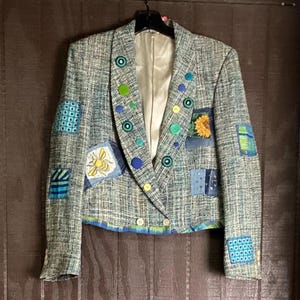 May include: A light blue tweed blazer with colorful patches and decorative buttons. The blazer features a variety of embroidered patches, including a bee, a sunflower, and geometric designs. The buttons are in shades of green, blue, and yellow.