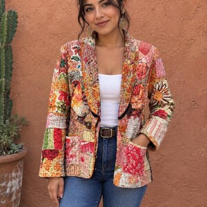 Könnte beinhalten: Ein gesteppter Blazer mit einem lebendigen Patchwork-Design aus verschiedenen Blumen- und geometrischen Mustern in Rot-, Orange-, Gelb- und Grüntönen. Die Jacke hat ein steigendes Revers, lange Ärmel und zwei aufgesetzte Vordertaschen mit schwarzer Kante.