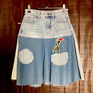 May include: A denim skirt with a patchwork design. The top portion is light blue denim, while the lower section is a darker blue. It features a striped pocket with a floral applique and a striped circle patch. The skirt is hanging on a metal hanger.