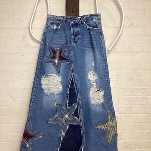 May include: A denim skirt with a distressed look, featuring star-shaped patches in plaid and patterned fabrics. The skirt has a front slit and is hung on a white, curved hanger.