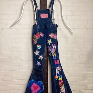 Può includere: Salopette in denim blu con gambe a zampa, decorate con toppe colorate tra cui stelle, fiori e un cuore tie-dye. La parola "HUMAN" è scritta su una gamba. La salopette è appesa a una gruccia bianca contro un muro di mattoni bianchi.