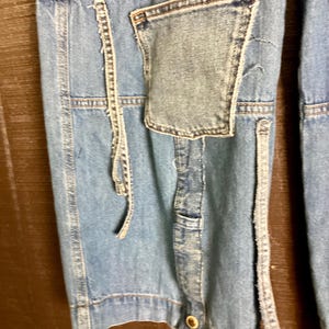 May include: Close-up of light blue denim pants with a patchwork pocket and decorative stitching. The pants have a button closure and a relaxed fit. The denim has a faded appearance, suggesting a vintage or worn style.