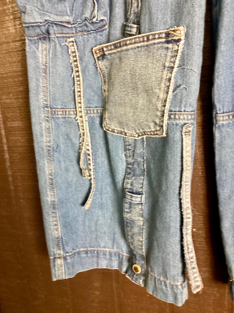 May include: Close-up of light blue denim pants with a patchwork pocket and decorative stitching. The pants have a button closure and a relaxed fit. The denim has a faded appearance, suggesting a vintage or worn style.